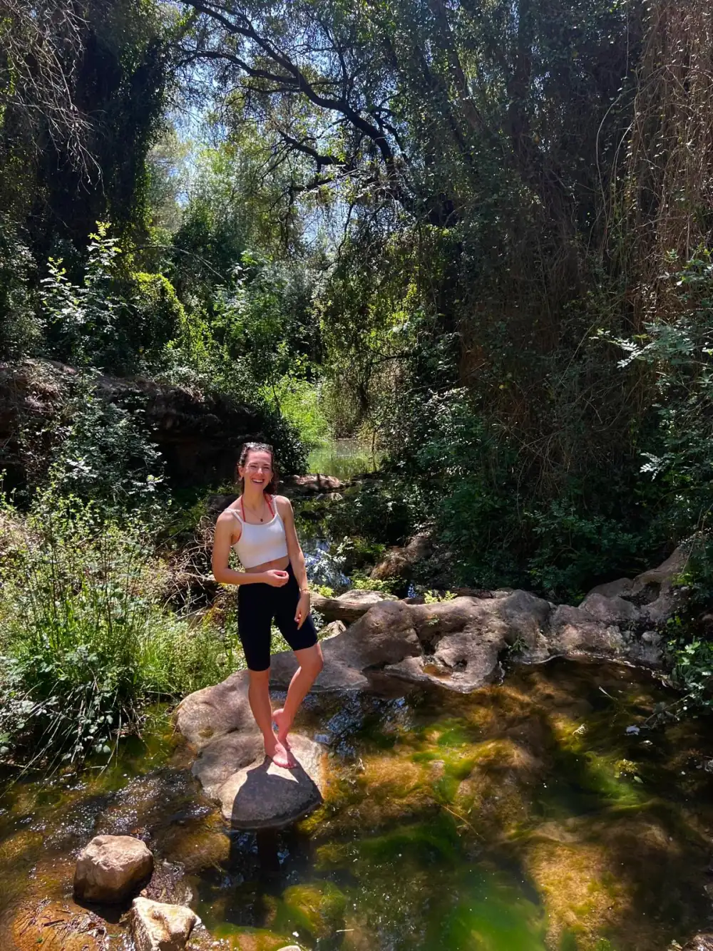 Chrissi balancing on a stone in a river, sunlight filtering through leaves
